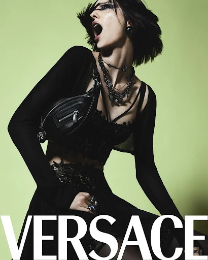 A woman with dark hair poses in a black outfit and jewelry in a studio with a lime green background. The Versace logo is visible.