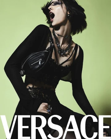 A woman with dark hair poses in a black outfit and jewelry in a studio with a lime green background. The Versace logo is visible.