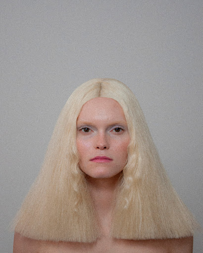 Close-up portrait of a fair-skinned adult female with a serious expression, heavy white eye makeup, and crimped blonde hair against a grey background.