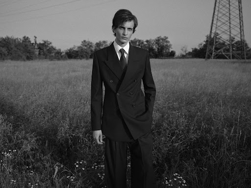 Monochrome portrait of a male model in a suit posing in a field with a power line structure in the background under natural light.