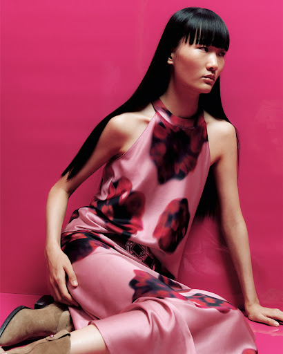 A stylish Asian woman in a floral dress poses against a vivid pink backdrop, showcasing fashion and beauty with a modern edge.