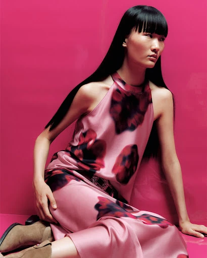A stylish Asian woman in a floral dress poses against a vivid pink backdrop, showcasing fashion and beauty with a modern edge.