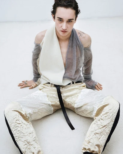 Young male fashion model sits on white floor wearing sheer top, textured pants and belt.  Fashion editorial with studio lighting.