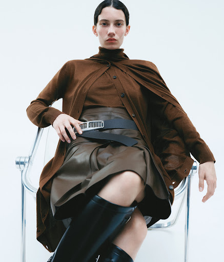 A woman poses while sitting in a chair in a studio wearing a brown sweater, skirt, and black boots against a white backdrop.