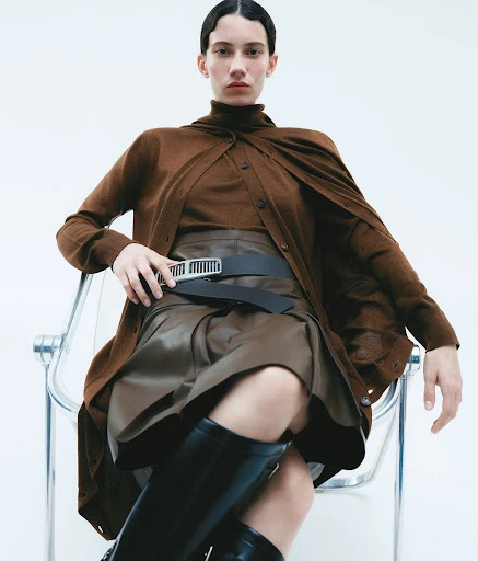 A woman poses while sitting in a chair in a studio wearing a brown sweater, skirt, and black boots against a white backdrop.