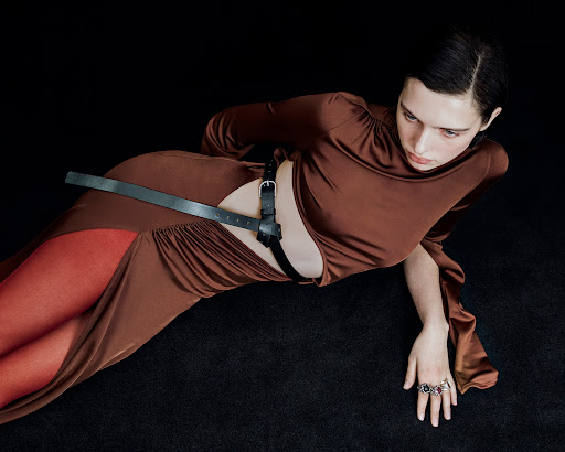 A woman poses in a brown dress and black harness against a dark backdrop, creating a high-fashion editorial image with dramatic lighting.