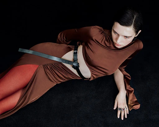 A woman poses in a brown dress and black harness against a dark backdrop, creating a high-fashion editorial image with dramatic lighting.