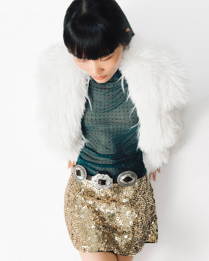 A woman with dark hair poses in a sheer green top, white faux fur jacket, and gold sequin skirt against a white backdrop.