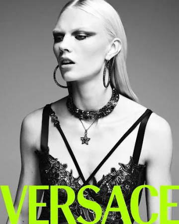Monochrome portrait of a fashion model wearing a black bra top and statement jewellery, shot against a gray background.