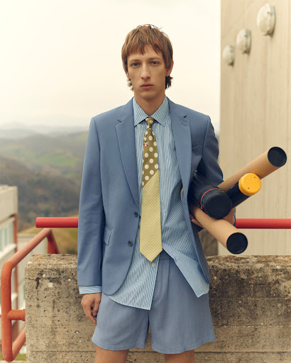 A fair-skinned male model in a blue suit and shorts holds rolled documents on a concrete balcony against a blurred landscape backdrop.