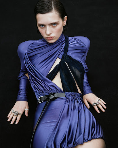 A young adult woman poses in a purple draped dress with a black belt and rings, set against a dark background in a studio shot.