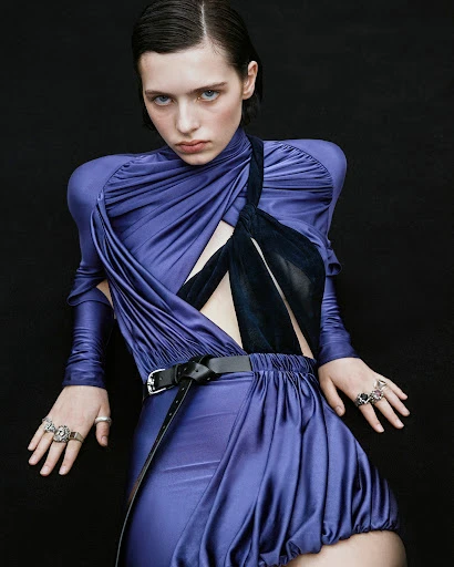 A young adult woman poses in a purple draped dress with a black belt and rings, set against a dark background in a studio shot.