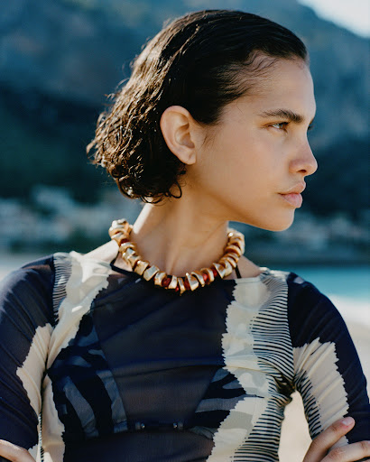 Profile shot of a woman with dark hair wearing a patterned top and a gold necklace outdoors on a beach with a mountainous background.