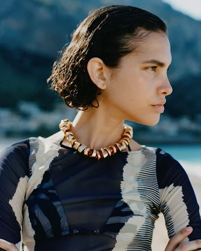 Profile shot of a woman with dark hair wearing a patterned top and a gold necklace outdoors on a beach with a mountainous background.
