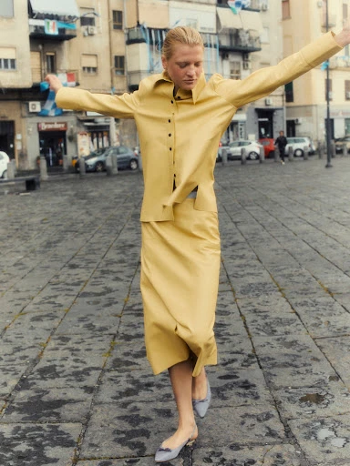 Woman in a yellow skirt suit dances with outstretched arms on a stone-paved city square, buildings in the background.