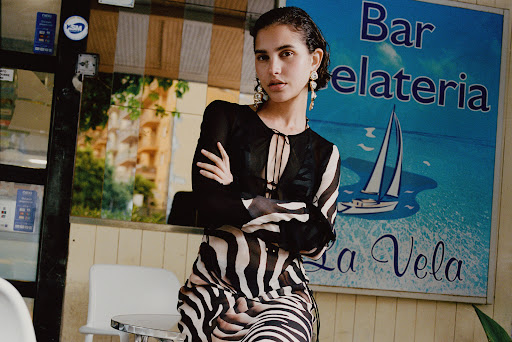 Adult woman in zebra-striped dress stands indoors near a gelato bar; she poses in front of blue sign with white sailboat graphic.