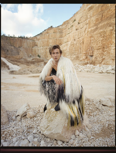 Fashionable young adult in dramatic fur coat and embellished top, crouching on rock in front of tall quarry under bright, cloudy sky.