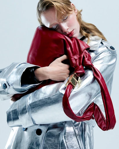 Fashion shot of a model embracing a red leather handbag while wearing a metallic silver trench coat in a bright studio setting.