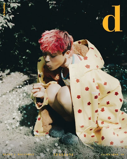 Young adult with pink hair sitting in grass wearing a yellow raincoat blowing a dandelion in an outdoor setting. Magazine cover.