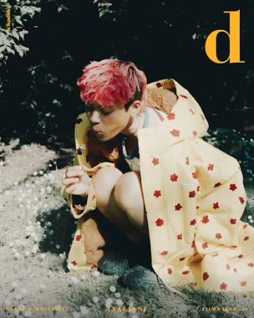 Young adult with pink hair sitting in grass wearing a yellow raincoat blowing a dandelion in an outdoor setting. Magazine cover.