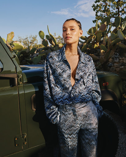 A woman in a patterned top and pants stands next to a green vehicle, posing in an outdoor setting with cactus plants and a blue sky.