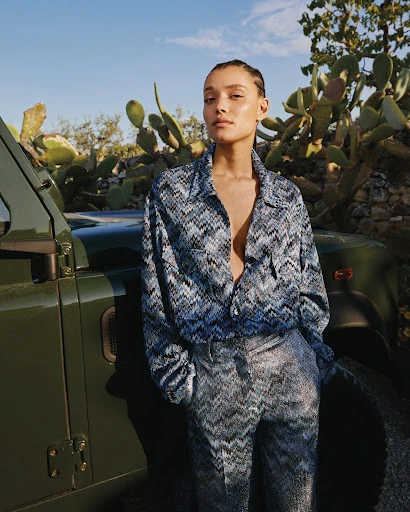 A woman in a patterned top and pants stands next to a green vehicle, posing in an outdoor setting with cactus plants and a blue sky.