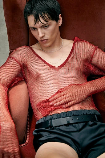 Portrait of a young adult male in a red fishnet top and black shorts sitting in a red chair looking downward in an indoor environment.