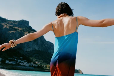 Model in blue gradient slip dress with arms outstretched against an ocean and coastal cliff background