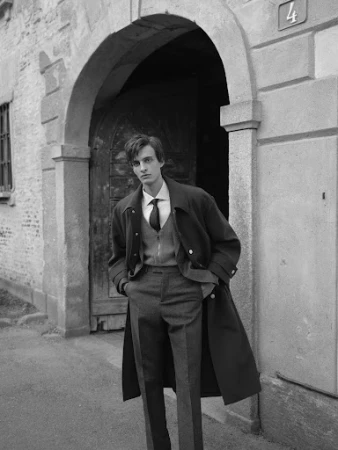 Monochrome portrait of a young man wearing a coat and tie standing in front of an arched doorway