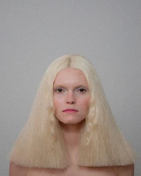 Close-up portrait of a woman with pale skin and long blonde crimped hair against a plain grey background