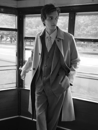 Monochrome portrait of a young male model wearing a trench coat, vest, and tie inside a transit vehicle