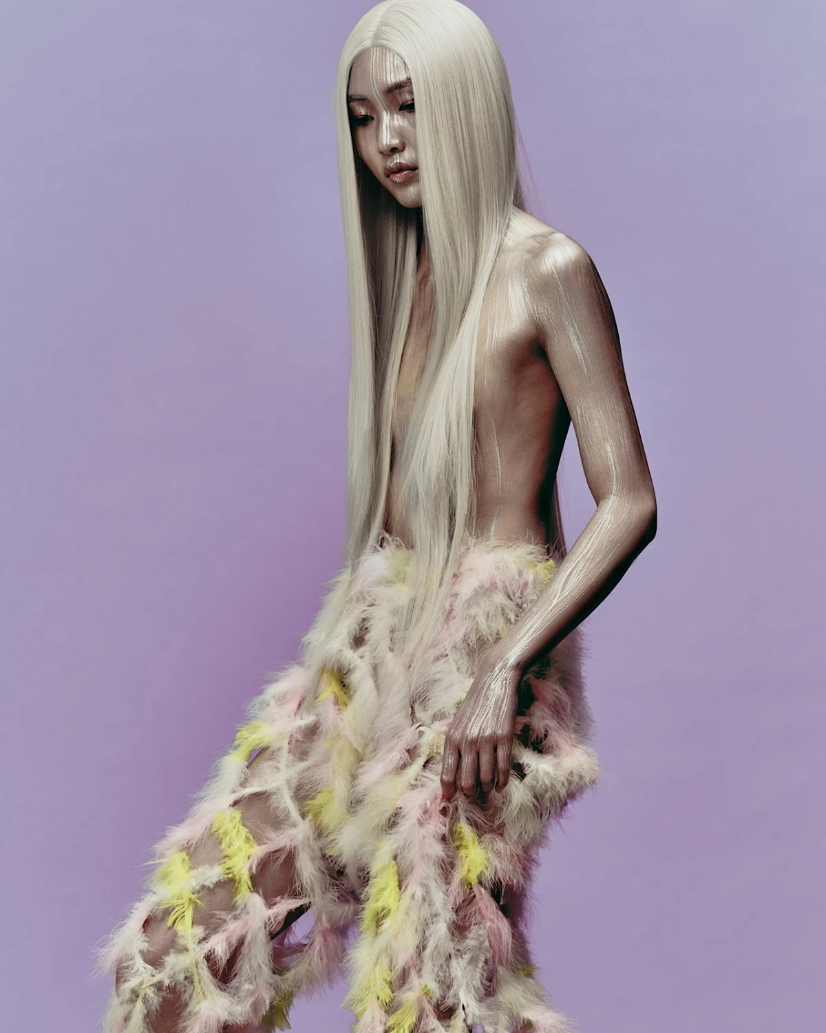 A person with pale skin and long, platinum-blonde hair poses in an artful, semi-nude manner, wearing feathered pants against a lavender backdrop.
