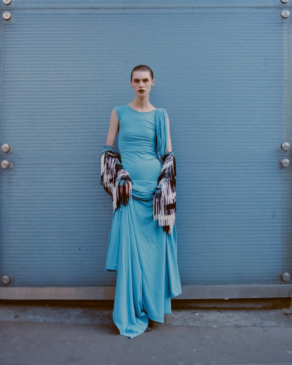 A woman in a long, light blue dress poses against a corrugated light blue wall, draped with a multicolored shawl or scarf.
