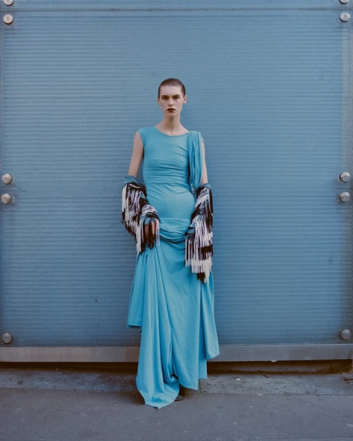 A woman in a long, light blue dress poses against a corrugated light blue wall, draped with a multicolored shawl or scarf.
