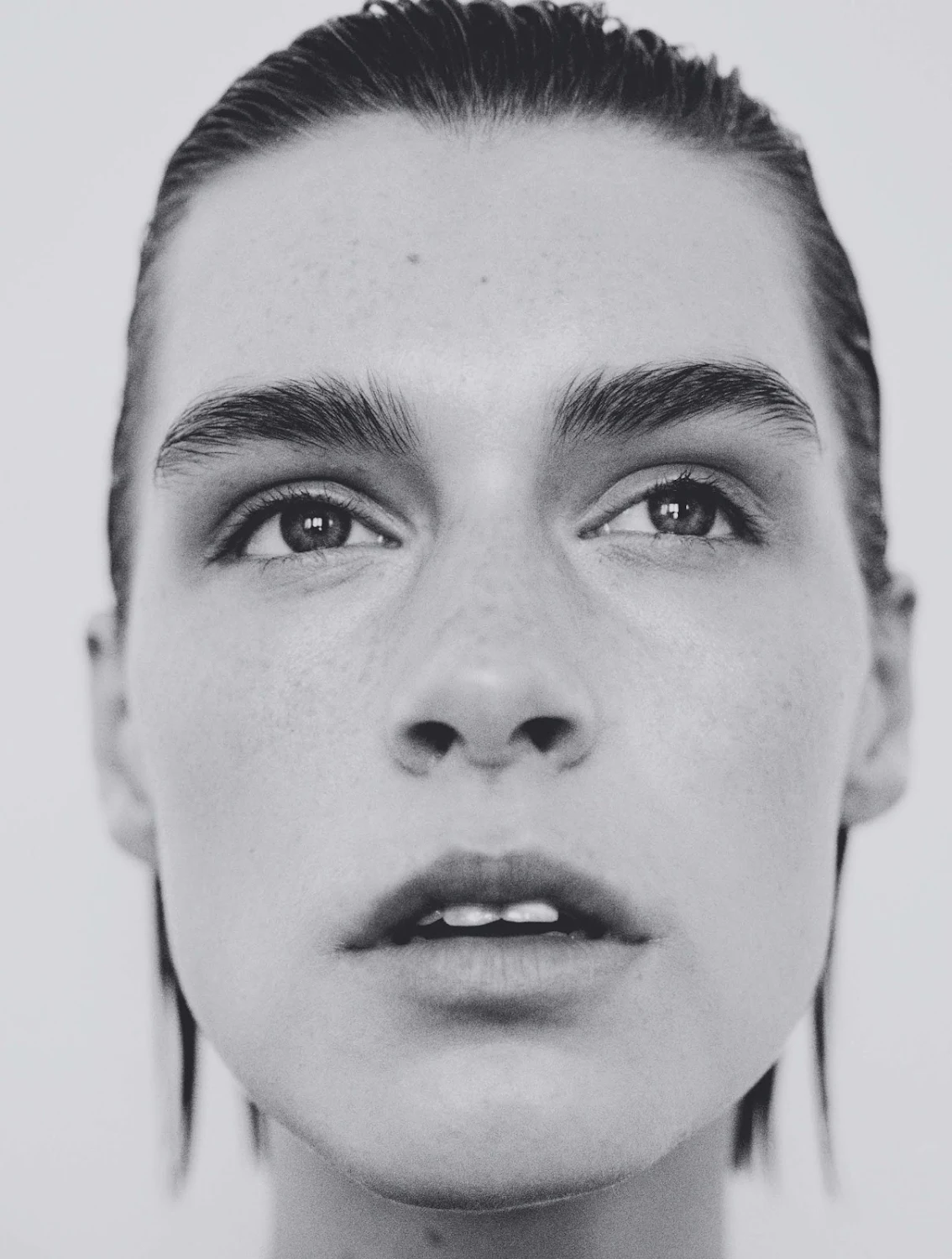 Close-up monochrome portrait of a model with slicked-back wet hair and natural makeup against a plain light background by Bastien Zorzetto.