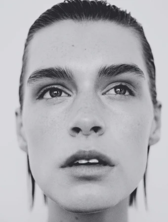 Close-up monochrome portrait of a model with slicked-back wet hair and natural makeup against a plain light background by Bastien Zorzetto.