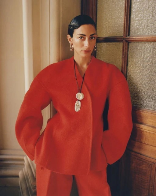 Adult woman in red suit poses indoors with geometric necklace and neutral backdrop by paneled door; natural light, minimalist fashion.