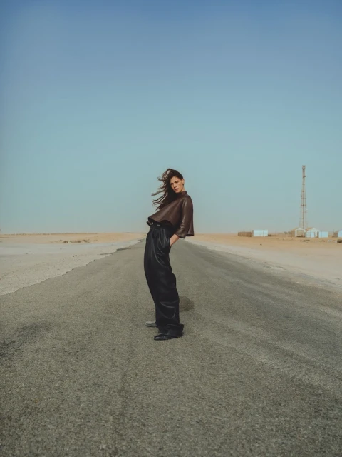 A woman in a brown top and black pants stands on a road in a desert landscape under a clear sky in a fashion editorial shot.