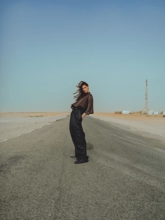 A woman in a brown top and black pants stands on a road in a desert landscape under a clear sky in a fashion editorial shot.