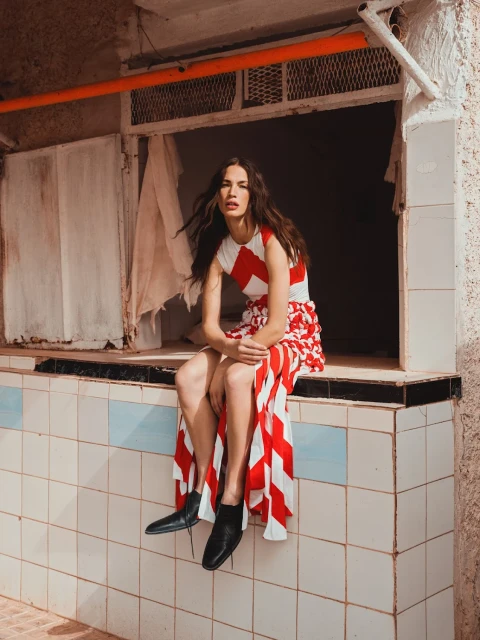 A woman in a red and white dress sits on a tiled ledge in an outdoor setting, wearing black boots with long hair and neutral expression.