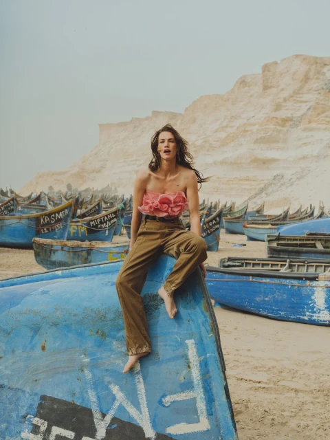 A woman wearing a floral top and brown pants sits on a blue boat on a sandy beach, with many similar boats and sandy bluffs in the background.