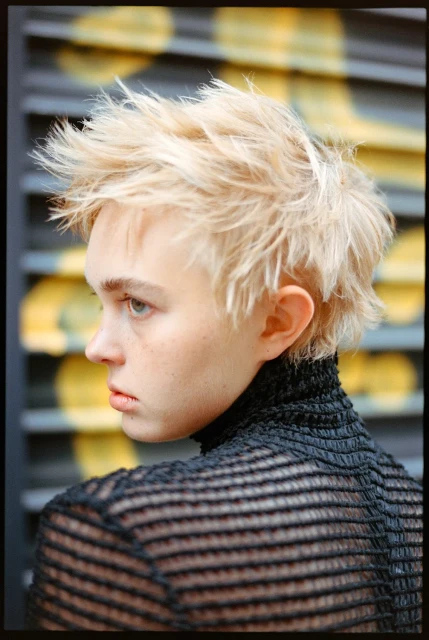 Portrait of a young adult with platinum blond spiky hair, wearing a black textured turtleneck shirt, against a gray and yellow backdrop.