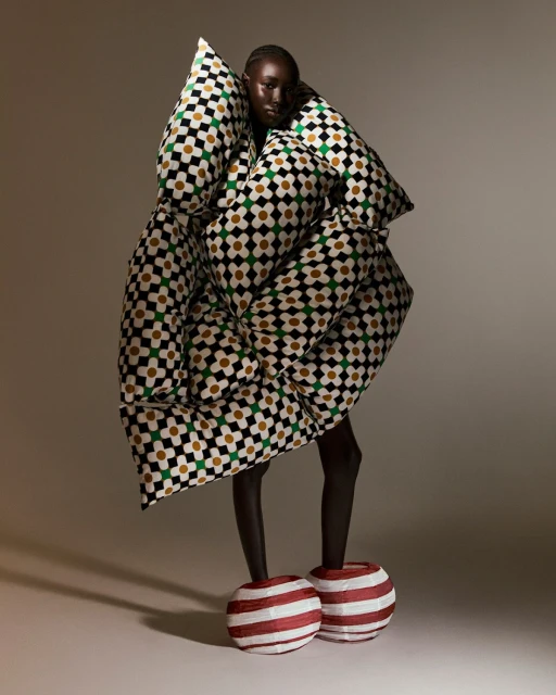 A fashion shot of a Black female model wearing an oversized patterned dress and striped spherical footwear in a brightly lit studio.