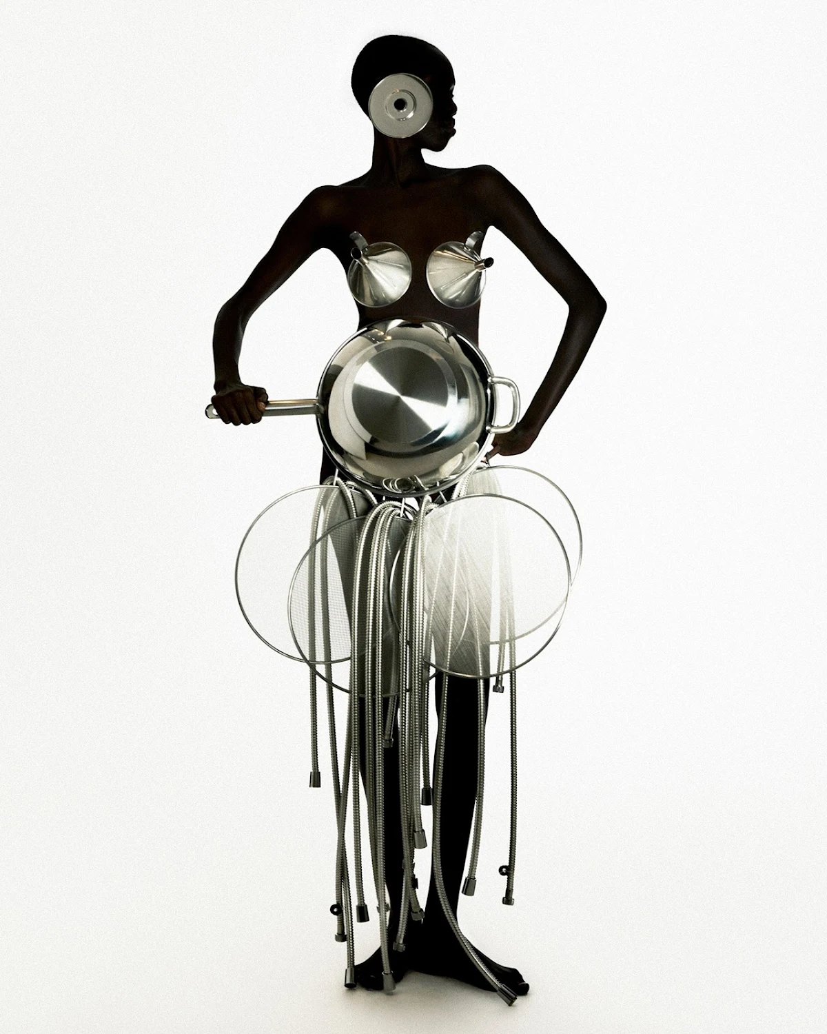 Model in avant-garde fashion ensemble with kitchenware elements, posing in a minimalist white studio. Silver cookware as clothing, artistic.