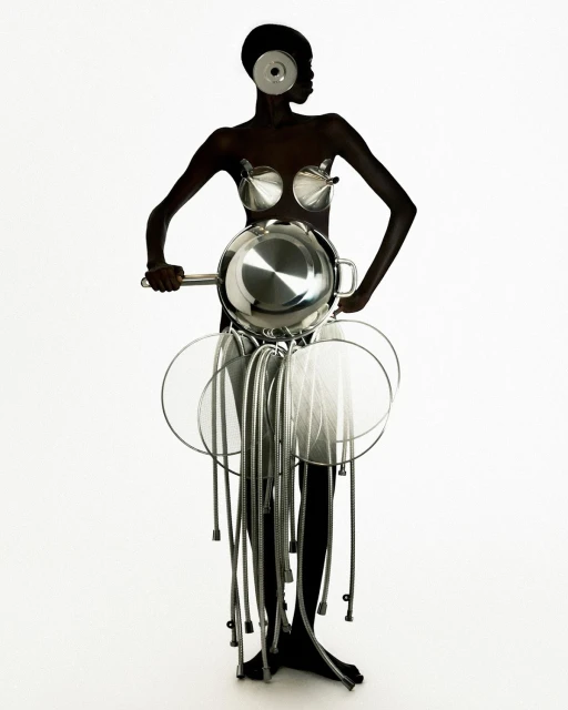 Model in avant-garde fashion ensemble with kitchenware elements, posing in a minimalist white studio. Silver cookware as clothing, artistic.