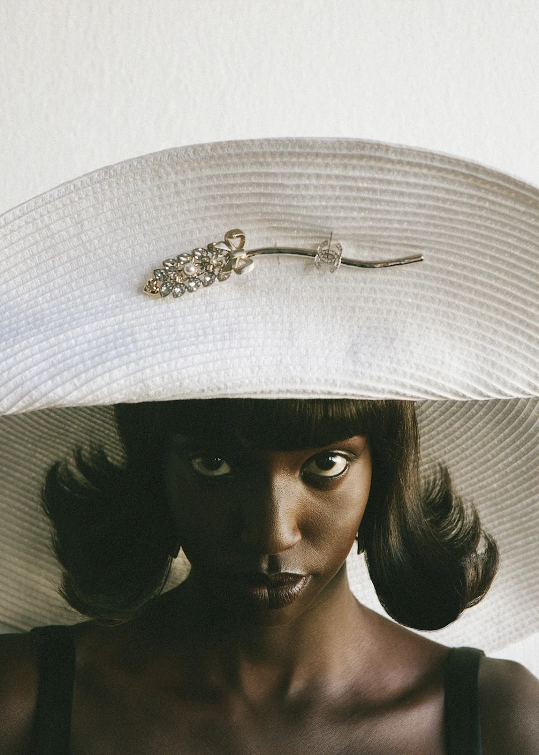 Close-up portrait of a Black female model wearing a large white hat and gazing directly at the camera in a brightly lit indoor setting.