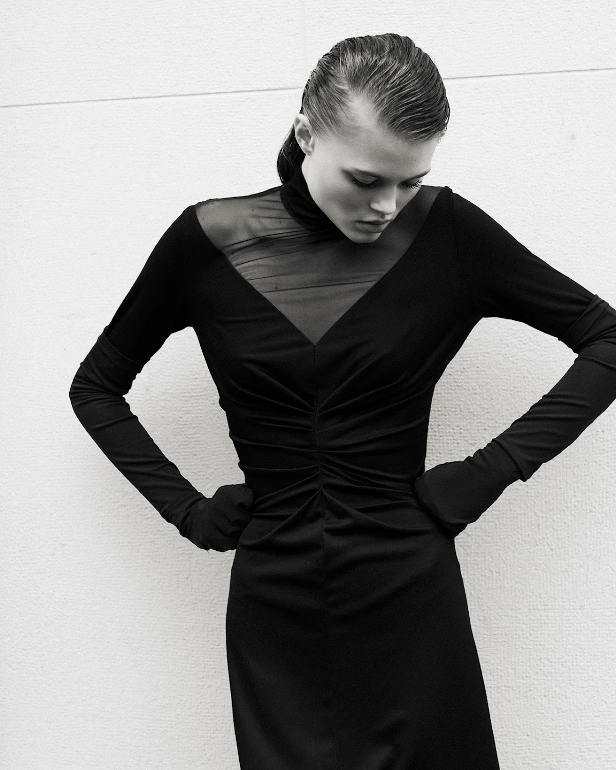 Black and white portrait of a fashion model with wet hair and a black, form-fitting dress posing against a plain white background.