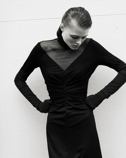 Black and white portrait of a fashion model with wet hair and a black, form-fitting dress posing against a plain white background.
