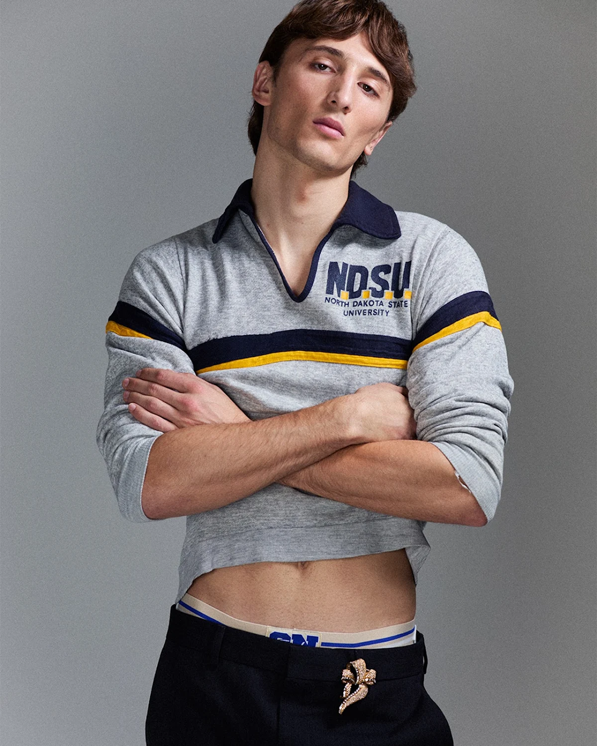 An adult man poses indoors, wearing a grey NDSU collared sweatshirt and black pants with a gold flower embellishment on the waistband.