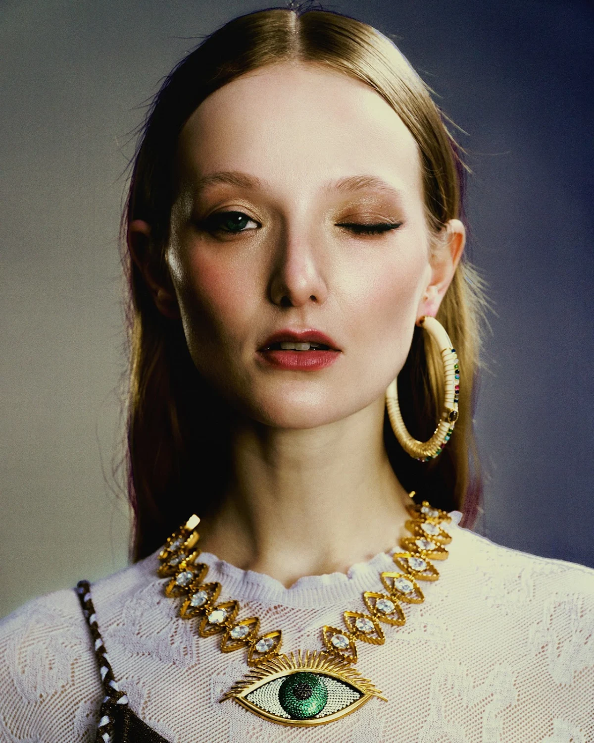Close-up portrait of a woman with auburn hair wearing a white sweater, large hoop earrings, and a distinctive eye-shaped gold necklace.